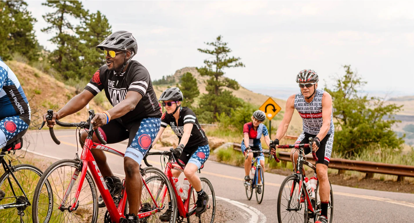 A group of cyclists in Phoenix-branded jerseys rides together on a winding road in a wooded landscape.